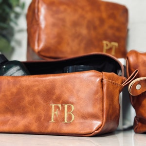 May include: Three brown leather toiletry bags with gold initials. The bag in the foreground has the initials "FB" embroidered on it. The bag in the middle has the initials "PT" embroidered on it. The bag in the background is closed.