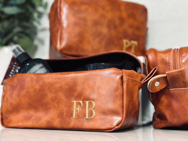 May include: Three brown leather toiletry bags with gold initials. The bag in the foreground has the initials "FB" embroidered on it. The bag in the middle has the initials "PT" embroidered on it. The bag in the background is closed.