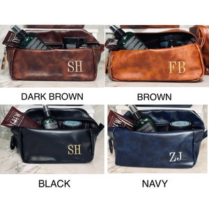 Op de afbeelding: Vier leren toilettassen in donkerbruin, bruin, zwart en marineblauw. Elke tas is rechthoekig met een ritssluiting en bevat verzorgingsproducten. De tassen zijn gepersonaliseerd met goud geborduurde initialen. De tekst geeft de kleur aan.