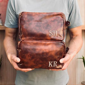 Op de afbeelding: Twee bruine leren toilettassen, op elkaar gestapeld, met gepersonaliseerde initialen "SH" en "KR" in wit geborduurd. De tassen hebben een vintage, distressed look en worden door een persoon vastgehouden. De tassen zijn rechthoekig met afgeronde hoeken en een klein handvat.