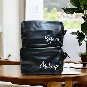 May include: Two black leather zippered cosmetic bags. The top bag has the name "Keyara" embroidered in white. The bottom bag has the word "Makeup" embroidered in white.