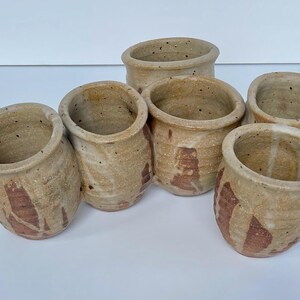 May include: Six small, handmade ceramic cups with a light brown glaze and brown speckles. The cups are arranged in a circle, with the rims facing up.