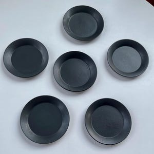 May include: A set of five circular, matte black plates arranged in a pentagonal formation. Each plate features a smooth, flat center surface with a slightly raised, rounded rim, providing a consistent and uniform appearance across all five pieces.