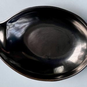 May include: A dark, oval-shaped ceramic dish with a small spout on one side. The dish has a glossy, reflective surface and a dark, metallic finish. The rim of the dish has a slightly lighter tone.