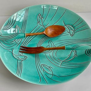 May include: A turquoise ceramic plate with a fish design, accompanied by a wooden spoon and fork. The spoon and fork have turquoise handles. The plate features black line art of fish and wavy lines.