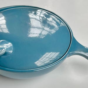 May include: A teal-colored ceramic pan with a lid and a handle. The lid has a unique double-loop handle. The pan is round with a curved handle extending from the side. The glossy finish reflects light.