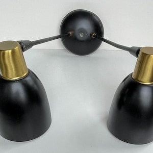 May include: A black wall sconce with two adjustable lights. Each light has a black shade and a gold-colored top. The sconce is mounted on a round black base and is ideal for a bedroom or living room.