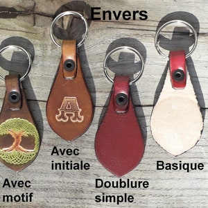 May include: A collection of leather keychains in various colors and designs. One keychain features a tree of life design, another has the letter "A", and others are solid colors. Each keychain has a metal ring and a leather loop with a rivet.