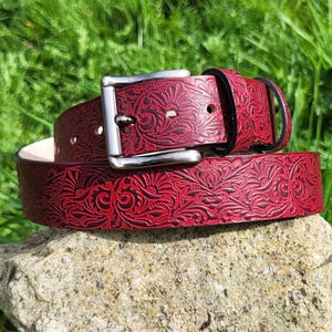 Peut inclure: Une ceinture en cuir rouge avec un motif floral en relief et une boucle argentée. La ceinture repose sur un rocher, avec de l'herbe verte en arrière-plan. La ceinture est de largeur moyenne.