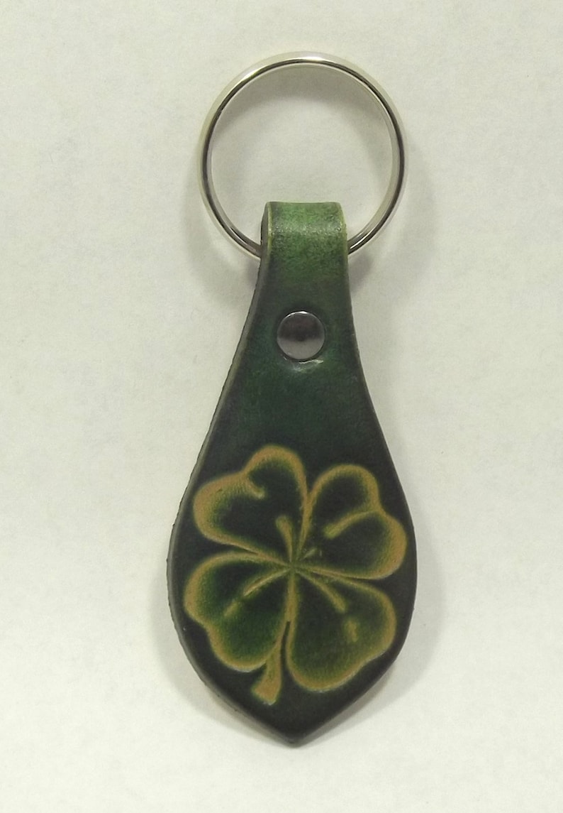 May include: Green leather keychain with a four-leaf clover design. The keychain features a silver metal ring and a dark green leather strap. The clover is embossed with lighter green and yellow details.