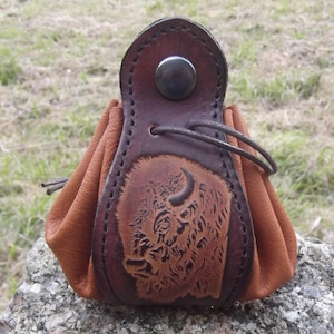 Może przedstawiać: Brązowa skórzana torebka na monety z tłoczonym wzorem głowy bawoła. Torebka ma sznurek do zamykania i metalowy guzik.