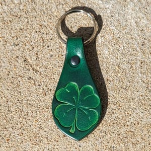 May include: A green leather keychain with a four-leaf clover design. The keychain has a metal ring and a black rivet. The clover is embossed on the leather.