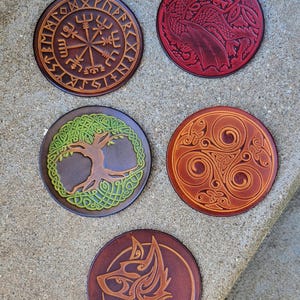Peut inclure: Cinq sous-verres ronds en cuir avec des motifs différents. Les sous-verres présentent un arbre de vie, un loup, un dragon, un nœud celtique et une boussole avec des runes.