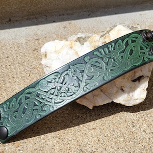 May include: Green leather bracelet with Celtic knotwork design. The bracelet features a detailed embossed pattern and is secured with two black snaps. The bracelet is displayed on a white rock.