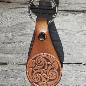 Peut inclure: Un porte-clés en cuir marron avec un motif de nœud celtique. Le porte-clés a un anneau de clé argenté et une lanière en cuir marron.