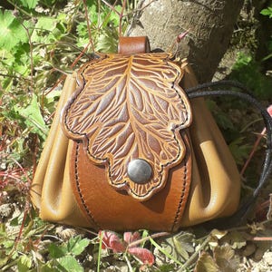 Könnte beinhalten: Eine braune Ledertasche mit einem Blattdesign und einem Metallknopfverschluss. Die Tasche hat einen Kordelzugverschluss und ist aus weichem, geschmeidigem Leder gefertigt.