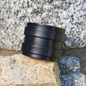 May include: A black leather cuff bracelet with multiple layers of leather. The bracelet is on a rock surface.