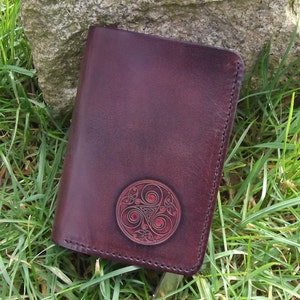 Peut inclure: Un portefeuille en cuir bordeaux avec un emblème circulaire décoratif. L'emblème présente un design de triskèle celtique de couleur brun rougeâtre. Le portefeuille rectangulaire est fermé, avec des coutures sur les bords.