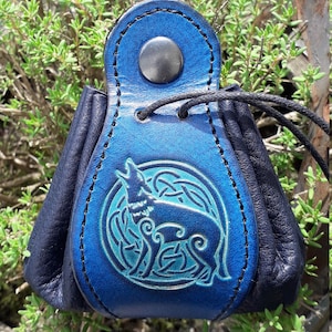 Op de afbeelding: Een klein blauw leren tasje met een zwarte koordsluiting. Het tasje heeft een Keltisch knoopontwerp met een wolf die naar de maan huilt. Het tasje is perfect voor het bewaren van kleine items zoals sieraden of munten.