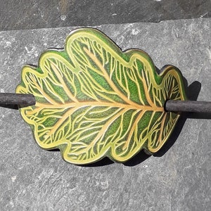 Puede incluir: Una pinza para el pelo de cuero verde y dorado en forma de hoja de roble con un palo de madera negro.