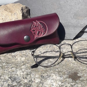 Peut inclure: Un étui à lunettes en cuir bordeaux avec un motif celtique décoratif et une fermeture à pression. L'étui est posé sur une surface en pierre grise à côté d'une paire de lunettes rondes à monture argentée.