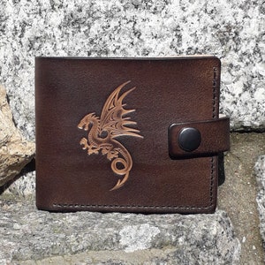 Peut inclure: Portefeuille en cuir marron avec un motif de dragon détaillé. Le portefeuille est doté d'une fermeture à bouton-pression et est fabriqué en cuir texturé. Le dragon est en relief sur le devant.
