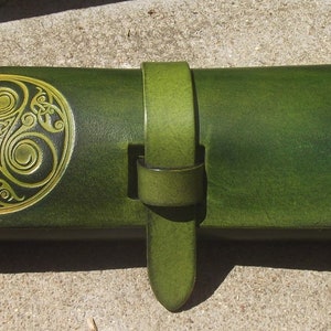 Pode incluir: Uma carteira de couro verde com um design de nó celta dourado e uma alça de couro verde.