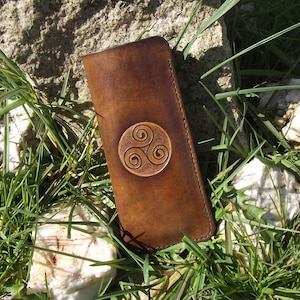 Könnte beinhalten: Eine braune Ledertasche mit einem keltischen Knotendesign. Die Tasche ist geschlossen und liegt auf einem Bett aus grünem Gras.
