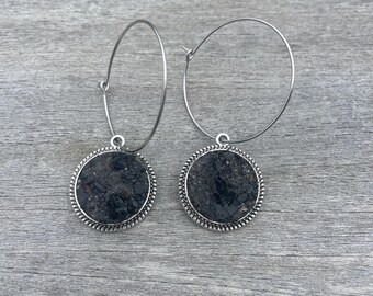 May include: A pair of hoop earrings with a circular, dark gray stone pendant. The stone is set in a silver-colored metal frame with a beaded edge. The earrings are displayed on a gray wooden surface.