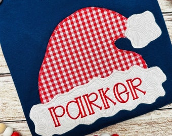 May include: A navy blue long-sleeved shirt with a red and white gingham Santa hat applique. The name "Parker" is embroidered in red on the hat.