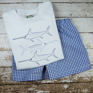 Pu&ograve; includere: Una maglietta bianca a maniche corte con tre pesci spada blu ricamati. La maglietta &egrave; abbinata a pantaloncini a quadri blu e bianchi.