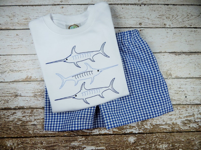 Pu&ograve; includere: Una maglietta bianca a maniche corte con tre pesci spada blu ricamati. La maglietta &egrave; abbinata a pantaloncini a quadri blu e bianchi.