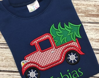 May include: A navy blue t-shirt with an embroidered red truck carrying a green Christmas tree. The name "Tobias" is embroidered in green below the truck.