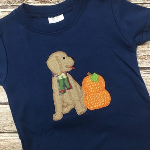 May include: A navy blue t-shirt with an embroidered design of a brown dog wearing a green and red scarf and standing next to a stack of two orange pumpkins.
