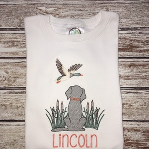 Peut inclure: T-shirt blanc brod&eacute; avec un chien noir et un canard volant au-dessus. Le chien est assis dans de hautes herbes et le texte "Lincoln" est brod&eacute; sous le chien.