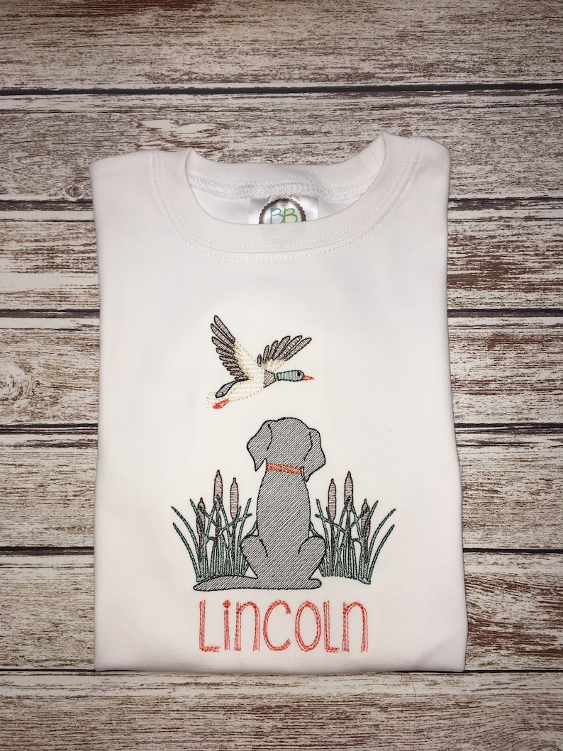 Peut inclure: T-shirt blanc brod&eacute; avec un chien noir et un canard volant au-dessus. Le chien est assis dans de hautes herbes et le texte "Lincoln" est brod&eacute; sous le chien.