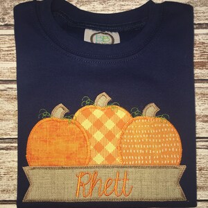 Peut inclure: Un t-shirt bleu marine avec un motif de trois citrouilles orange aux motifs diff&eacute;rents. Les citrouilles sont cousues sur une banni&egrave;re brune avec le nom "Rhett" brod&eacute; en fil orange.