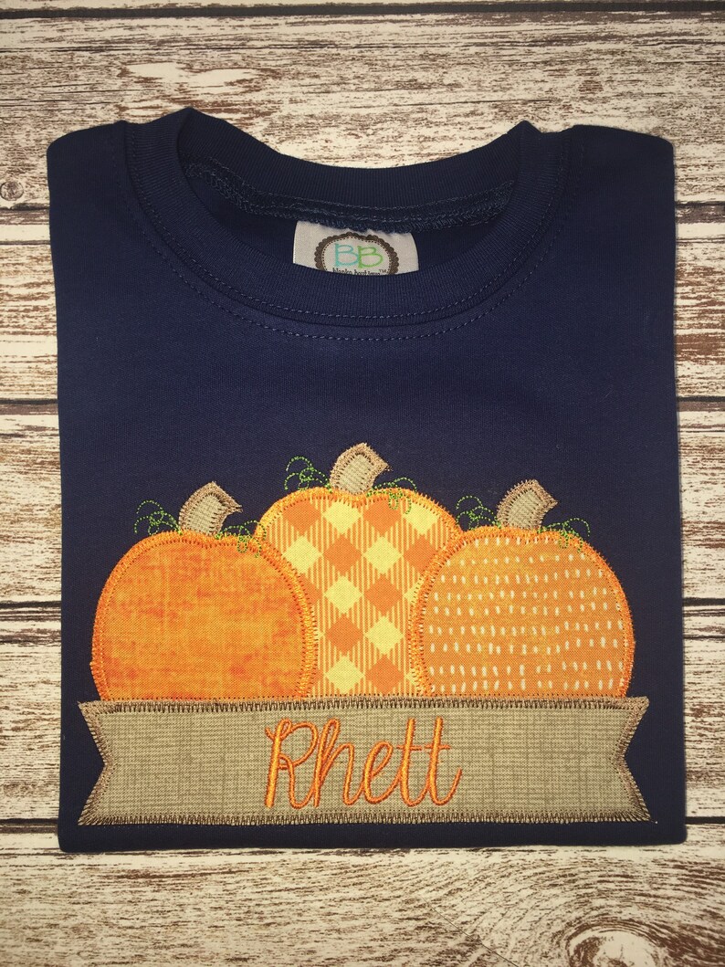 Peut inclure: Un t-shirt bleu marine avec un motif de trois citrouilles orange aux motifs diff&eacute;rents. Les citrouilles sont cousues sur une banni&egrave;re brune avec le nom "Rhett" brod&eacute; en fil orange.