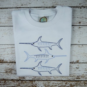 Pu&ograve; includere: Maglietta bianca con tre pesci spada ricamati in blu e bianco. I pesci spada sono disposti in linea verticale, con il pesce spada pi&ugrave; grande in alto e il pesce spada pi&ugrave; piccolo in basso.