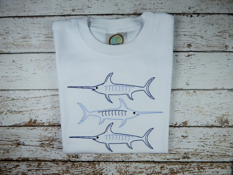 Pu&ograve; includere: Maglietta bianca con tre pesci spada ricamati in blu e bianco. I pesci spada sono disposti in linea verticale, con il pesce spada pi&ugrave; grande in alto e il pesce spada pi&ugrave; piccolo in basso.