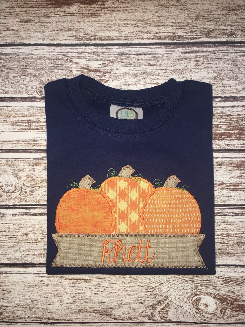 Peut inclure: Un t-shirt &agrave; manches longues bleu marine avec une banni&egrave;re personnalis&eacute;e et trois citrouilles brod&eacute;es. La banni&egrave;re indique "Rhett".