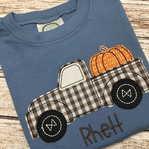 May include: A blue t-shirt with a brown and white checkered truck carrying an orange pumpkin. The truck has black wheels and the name "Rhett" is embroidered on the shirt.