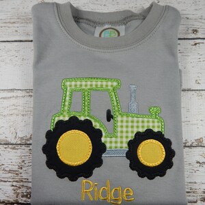 Puede incluir: Una camiseta gris con un tractor bordado en vichy verde y blanco, con ruedas negras y centros amarillos. El nombre "Ridge" est&aacute; bordado debajo del tractor.