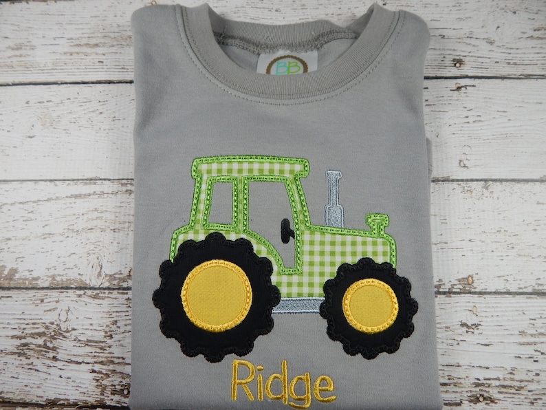 Puede incluir: Una camiseta gris con un tractor bordado en vichy verde y blanco, con ruedas negras y centros amarillos. El nombre "Ridge" est&aacute; bordado debajo del tractor.