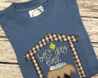 May include: A blue t-shirt with a brown and white checkered design of a manger with a baby inside. The text "best day ever" is embroidered on the manger.