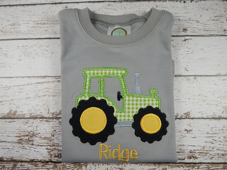 Puede incluir: Una camiseta gris con un dise&ntilde;o de tractor a cuadros verde y blanco y el nombre "Ridge" bordado en la parte inferior.
