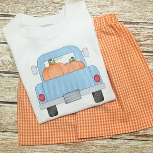 May include: A white short-sleeved shirt with an embroidered design of a blue pickup truck carrying three orange pumpkins. The shirt is paired with orange and white gingham shorts.
