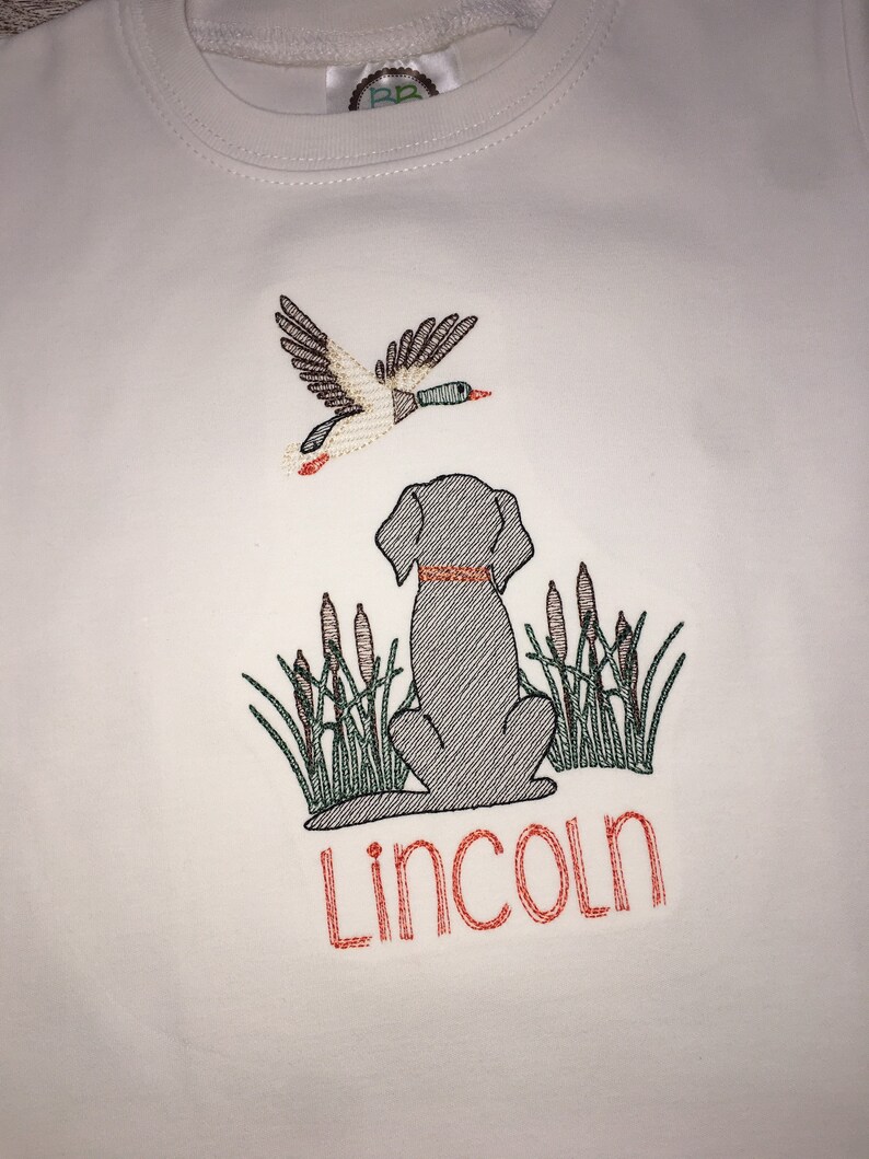 Peut inclure: Chemise blanche &agrave; manches longues avec un motif brod&eacute; repr&eacute;sentant un chien assis dans de hautes herbes avec un canard volant au-dessus. Le nom "Lincoln" est brod&eacute; sous le chien.