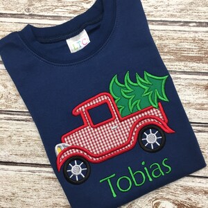 May include: A navy blue t-shirt with a red and white gingham truck carrying a green Christmas tree. The name "Tobias" is embroidered in green below the truck.