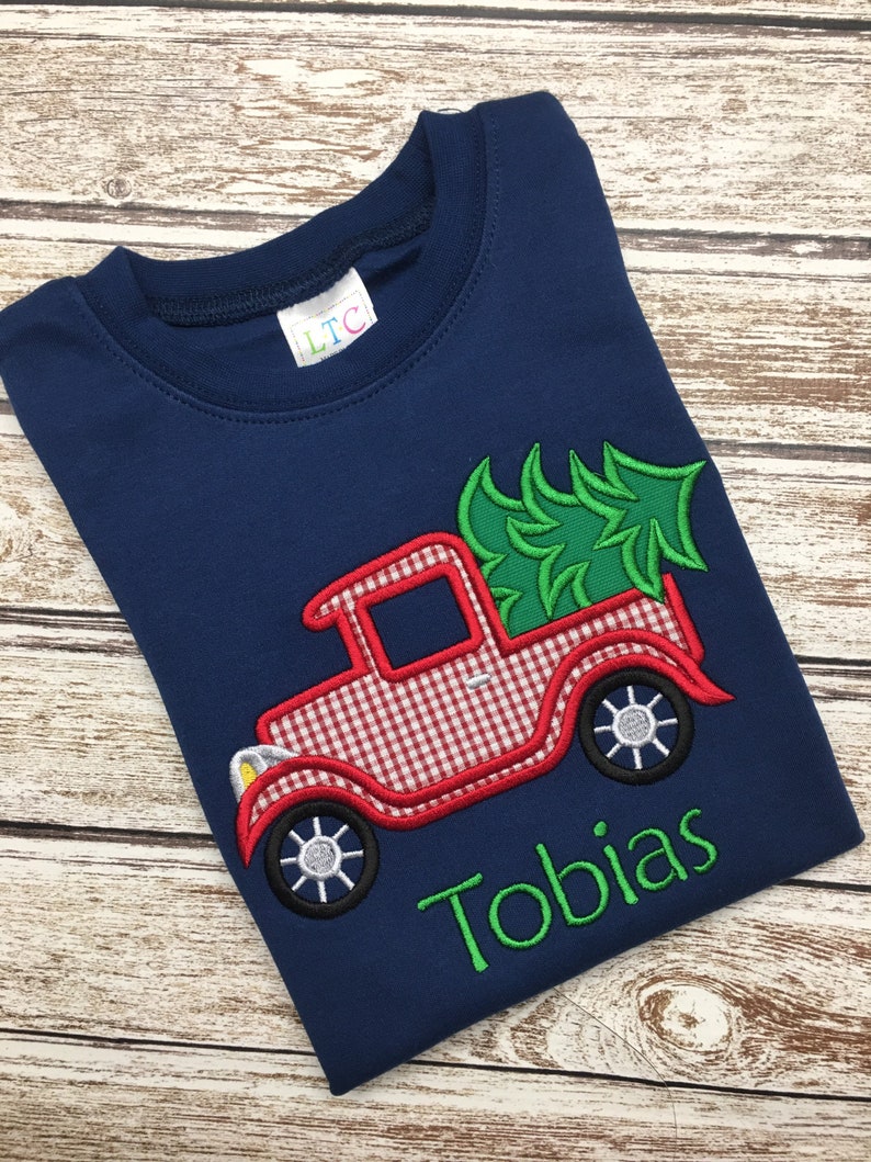 May include: A navy blue t-shirt with a red and white gingham truck carrying a green Christmas tree. The name "Tobias" is embroidered in green below the truck.